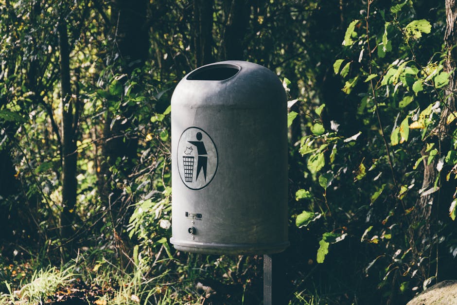 A cylindrical metal rubbish bin with a rounded top and large slot opening, situated outdoors against a backdrop of dense green foliage. The bin has a smooth, slightly reflective surface with a painted symbol of a person discarding waste into a bin on its front, indicating its purpose. It is mounted on a sturdy post, positioned on a patch of ground with grass and small plants nearby. The surrounding environment includes leafy shrubs and trees, with natural light filtering through the branches, casting soft shadows. This scene depicts an outdoor waste disposal point, which could be part of a private or public area where rubbish is responsibly collected, aligning with services provided by Waste Removal Surrey for local rubbish or waste management needs.