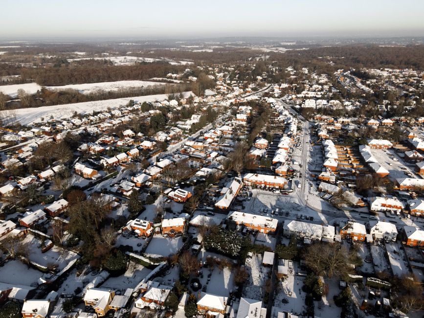 An aerial view of a residential neighbourhood during winter, showing rows of houses with snow-covered roofs, narrow streets lined with parked cars, and scattered trees. The houses are primarily brick buildings with pitched roofs, some with white or light grey roofing materials, and a mix of single and semi-detached layouts. The streets wind through the area, with snow visible on the driveways and pavements. In the background, open fields and patches of woodland extend into the distance, also lightly coated with snow. The sky is clear, providing bright natural lighting, and the overall scene reflects a quiet, winter day typical of suburban Surrey. This image illustrates a quiet suburban setting that could benefit from private waste collection or rubbish removal services, such as those offered by Waste Removal Surrey, to handle household waste outside of local authority collection.