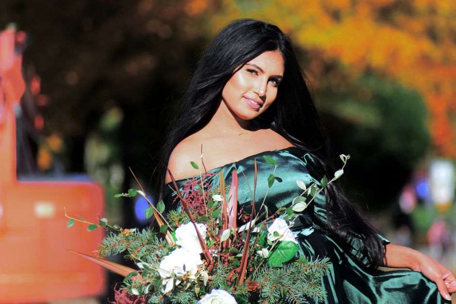 A young woman with long, dark hair and fair skin is standing outdoors in a park or garden setting during autumn. She is wearing an off-the-shoulder dark green satin dress. She is holding a large bouquet of various flowers and greenery, including white blooms, green leaves, and brown and reddish accents, which are arranged loosely and extend outward in different directions. In the background, blurred trees with orange foliage and a hint of a pathway or open space are visible, suggesting a natural environment. The lighting appears natural, highlighting her facial features and the textures of her dress and bouquet. This scene relates to private or on-site garden waste clearance activities, emphasizing the natural context of waste removal from garden or outdoor spaces, as provided by Waste Removal Surrey.