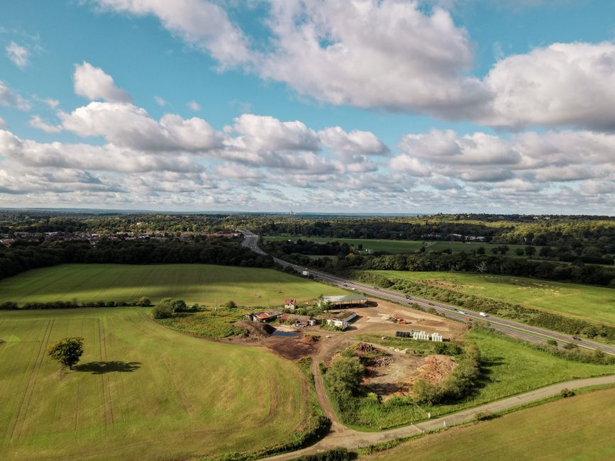 An aerial view of a rural landscape showing open green fields with a few scattered trees and a small construction site in the foreground, where earth has been excavated and some construction materials are visible. A narrow dirt path runs through the site, connecting it to the surrounding grassy area. In the middle distance, there is a dual carriageway with moving vehicles, bordered by hedgerows and trees. Beyond the road, additional fields and wooded areas stretch towards the horizon under a bright blue sky filled with fluffy white clouds. The scene suggests a mix of natural and semi-developed land, with evidence of land clearing that could relate to private waste or material removal services, such as those offered by Waste Removal Surrey, for on-site clearance or alternative waste handling at rural or semi-urban locations.