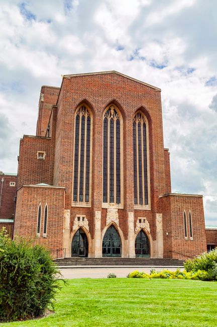 A large, historic church building constructed from red brick with tall, narrow Gothic-style windows featuring pointed arches and stained glass. The façade is symmetrical, with three arched doorways at ground level leading to the entrance, and decorative stone detailing around the doorways. The church is set on a slightly raised platform accessed by a short flight of steps. Surrounding the building is a well-maintained lawn with small yellow flowering plants and green bushes at the front. The sky above is partly cloudy with patches of blue visible, creating a natural lighting effect that highlights the textured brickwork and architecture of the church. The scene overall reflects a traditional, ecclesiastical structure, showing no signs of rubbish or waste, and emphasizing the building's historical and architectural significance, which may be relevant in the context of alternative waste handling or site management for historic locations.
