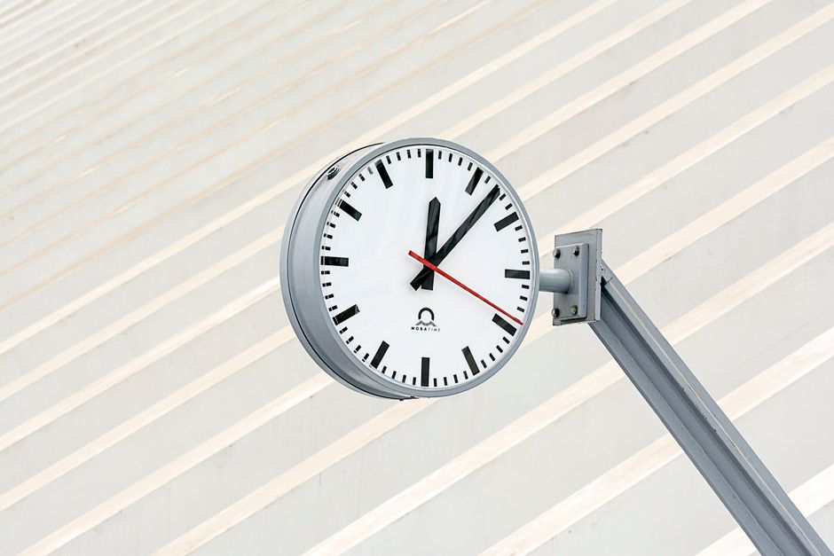 A large round wall clock with a white face, black hour and minute hands, and a red second hand, mounted on a metal arm extending from a wall with horizontal, off-white panels. The clock displays the time as approximately 11:55, and the environment appears to be a modern, indoor or outdoor public space with clean, lined surfaces. The clock is branded with a logo that suggests a connection to public or commercial settings, which may relate to scheduled waste collections or operational timings. The overall scene emphasizes clarity and precision, reflecting organizational aspects of waste management services offered by Waste Removal Surrey, especially in contexts where fixed schedules or independent waste disposal options are relevant to rubbish removal in Surrey.